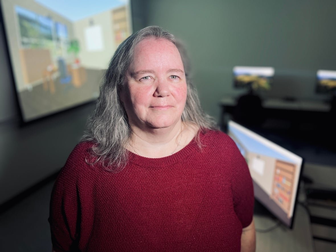 A portrait of a woman in a red sweater standing in a room with computers and a projection screen behind her.