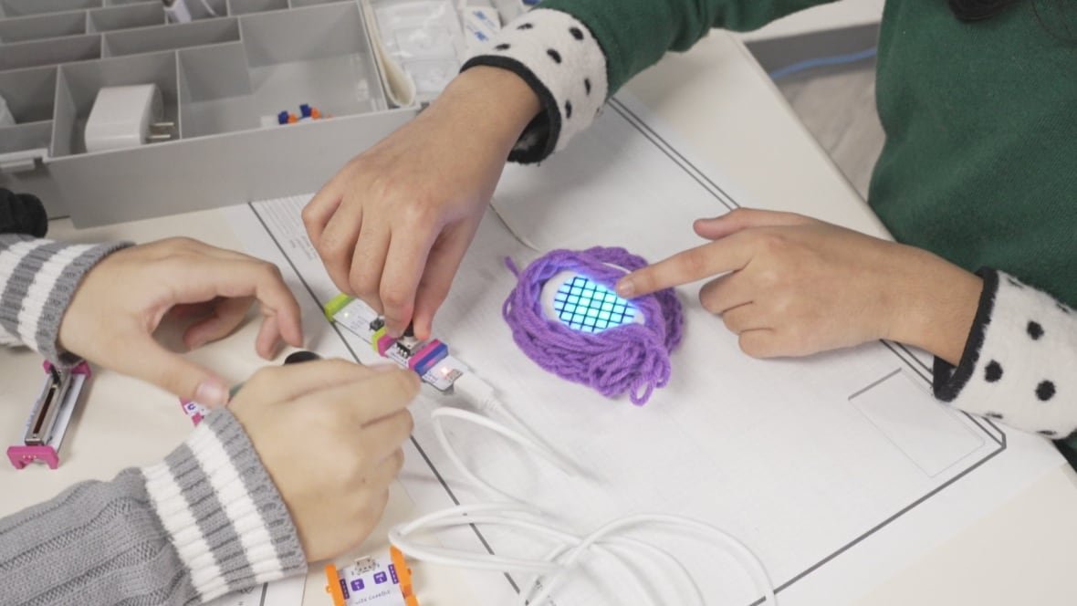 Close-up image on the hands of two girls as they assemble pieces of a simple circuit linked to a circular LED light with purple yarn wrapped around it.