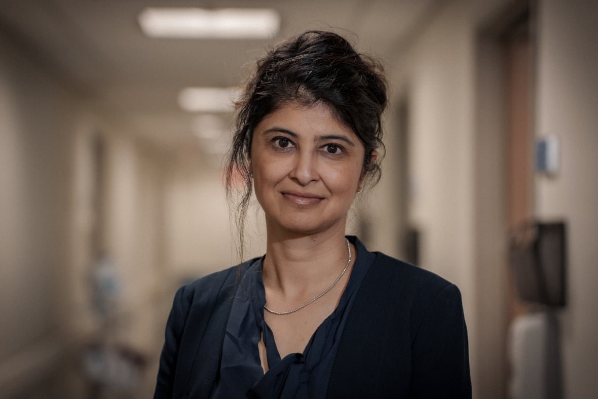 A woman with her hair back and wearing a black blouse and blazer stares at the camera in a hospital hallway. 