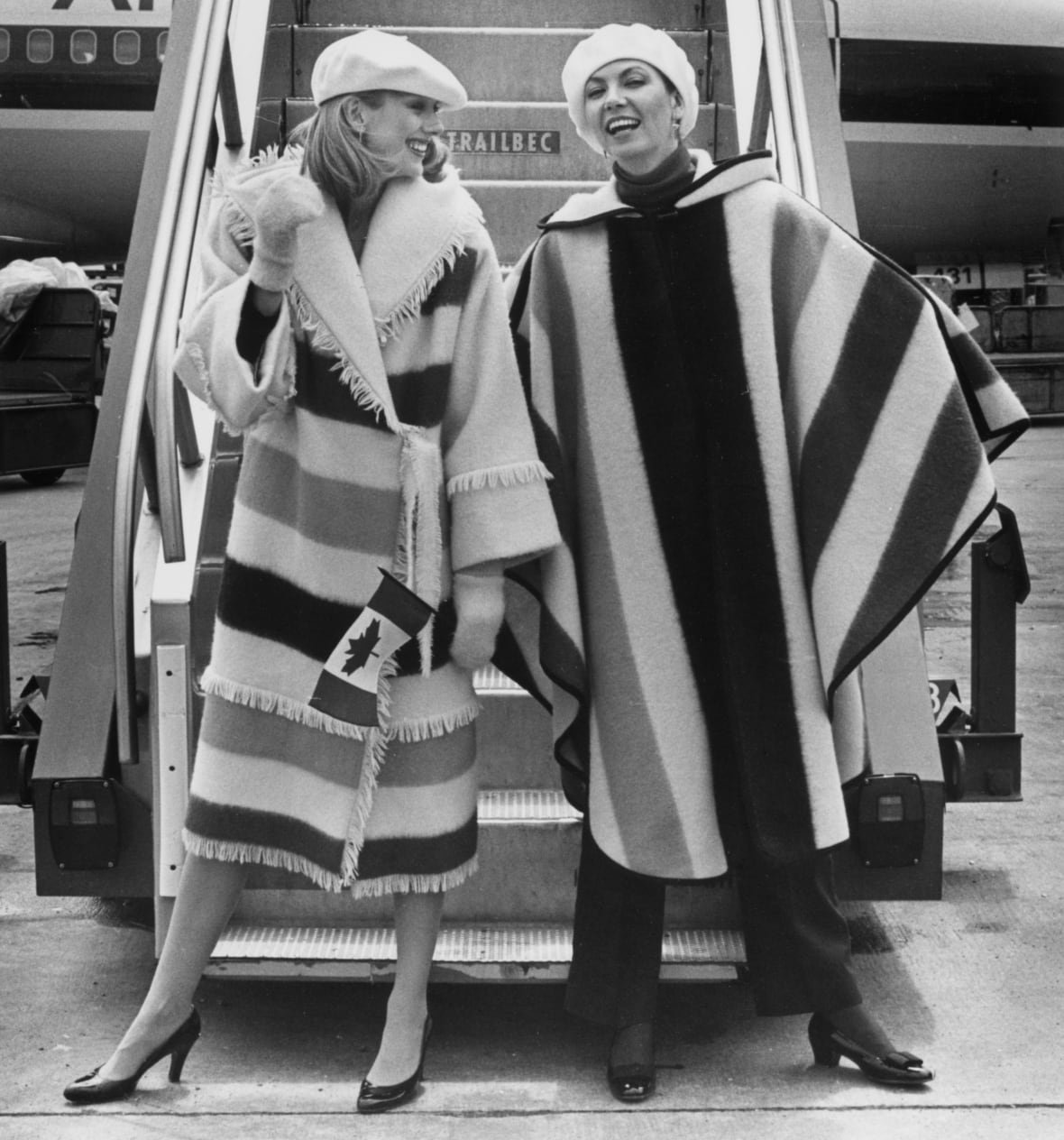 Two women wearing long coats made of striped fabric pose at the base of a staircase leading to a jetliner. 