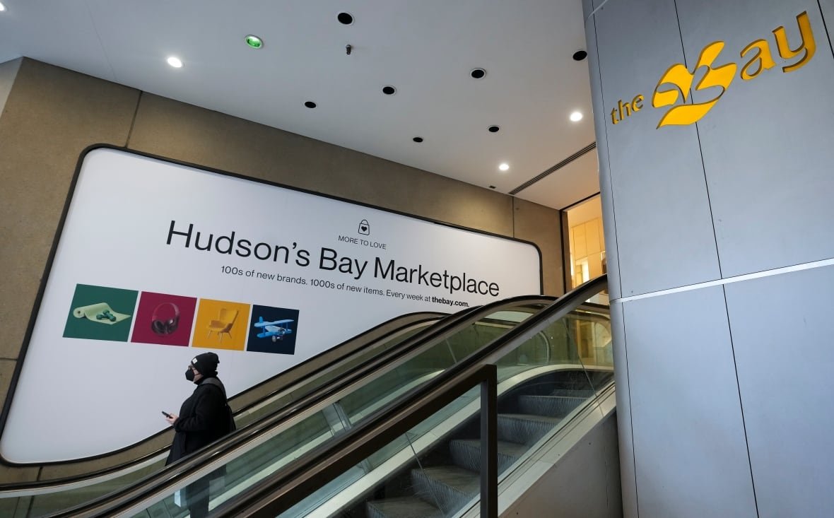 A person rides an escalator past a large sign reading 'Hudson's Bay Marketplace'
