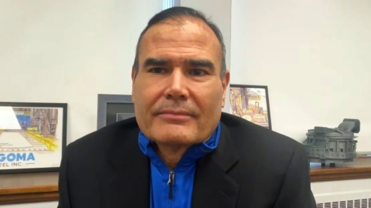 A man in a blue sweater and grey jacket looks into the camera from his office.