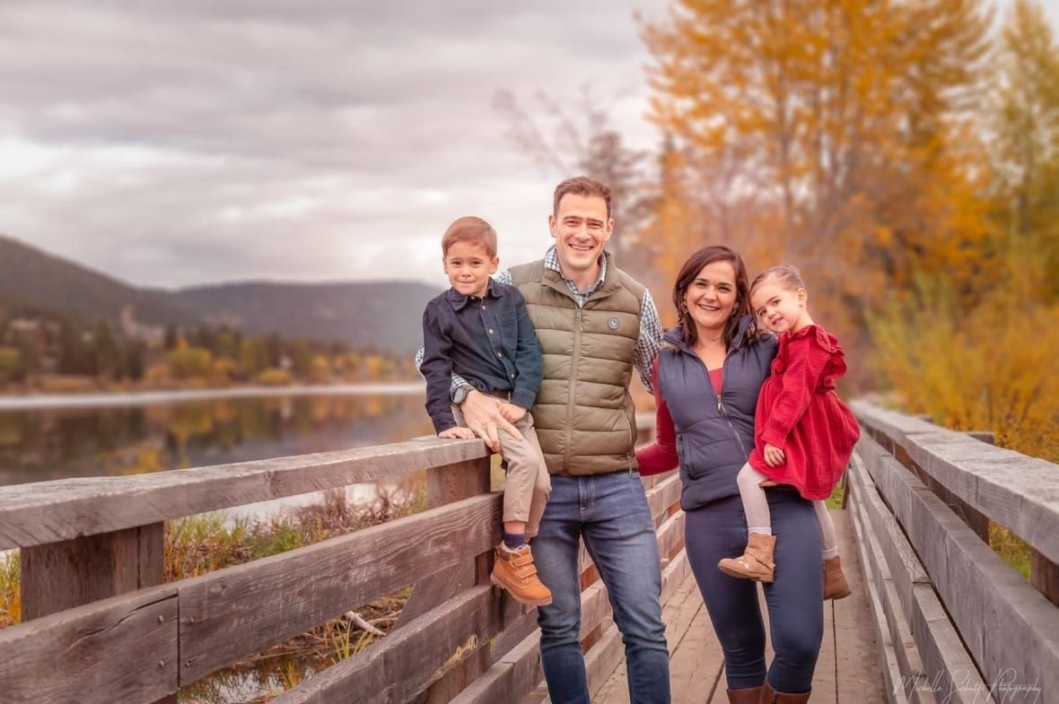 A family of four stands on a wooden bridge with fall leaves behind them.