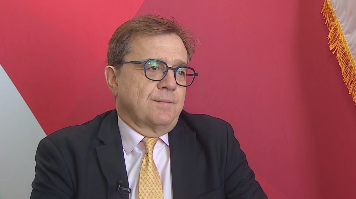 A politician sits in front of a red and white background.