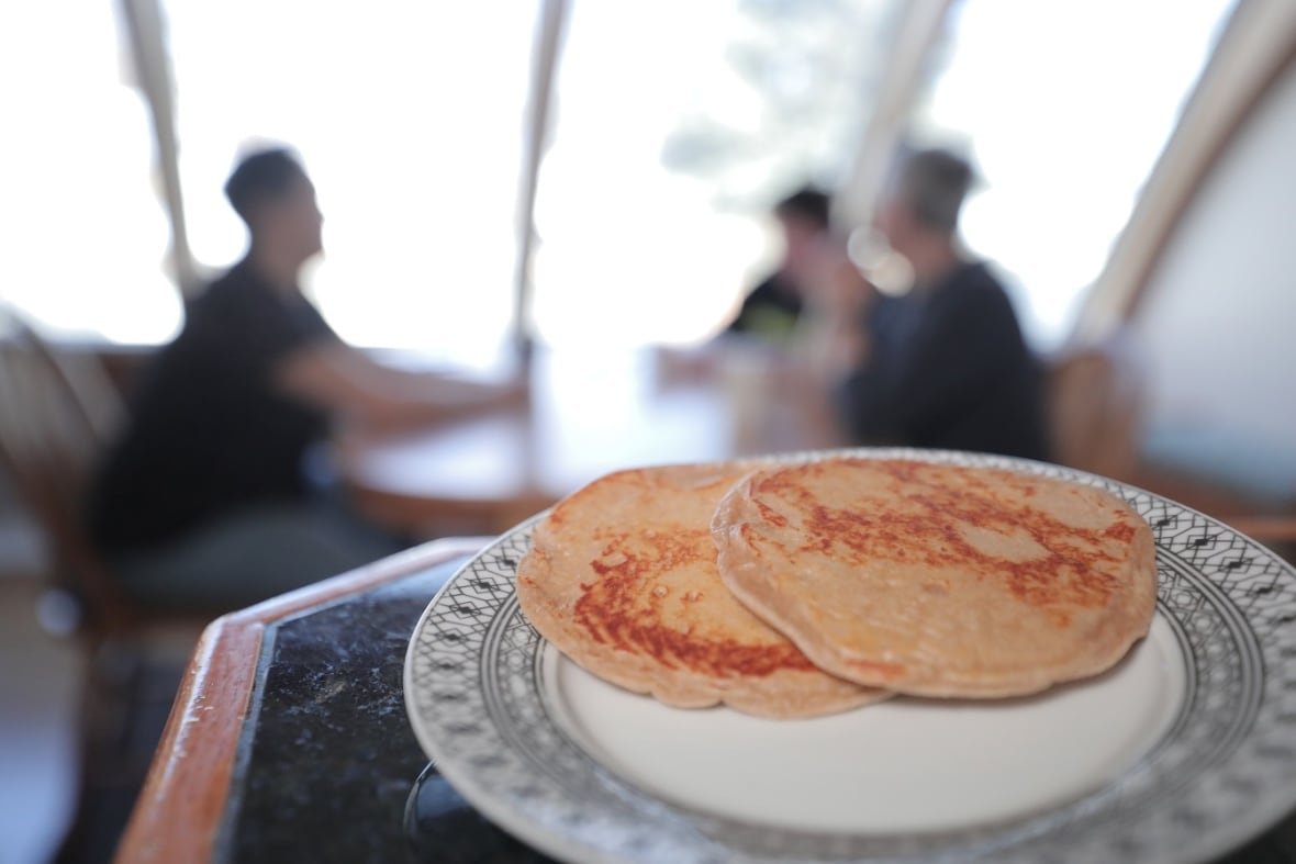 A plate of pancakes sits in focus in the foreground. In the background, three figures sit at a table, out of focus to the point that they can't be identified visually.