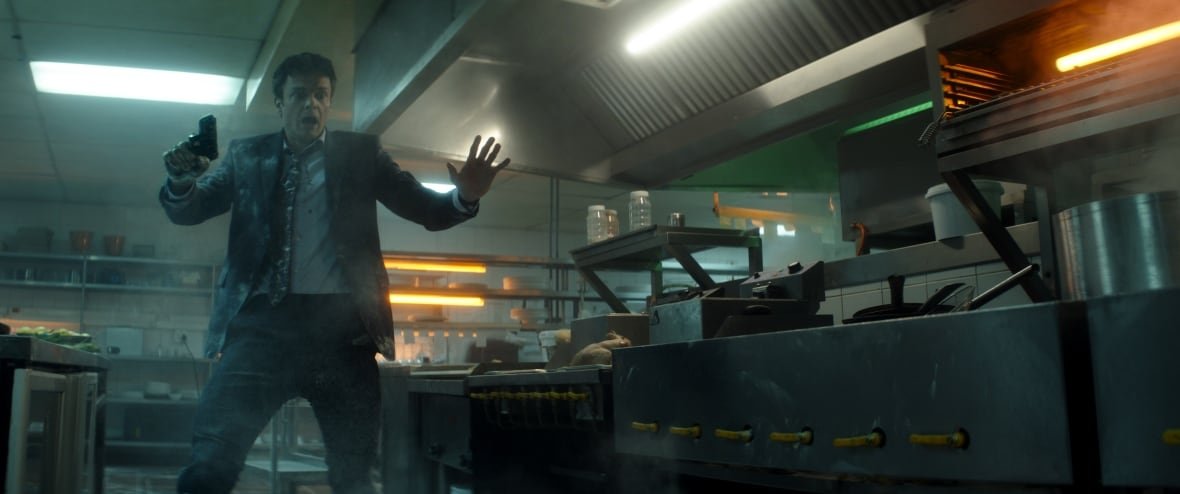 A frightened man in a commercial kitchen holds a gun to the side while holding his other palm out.