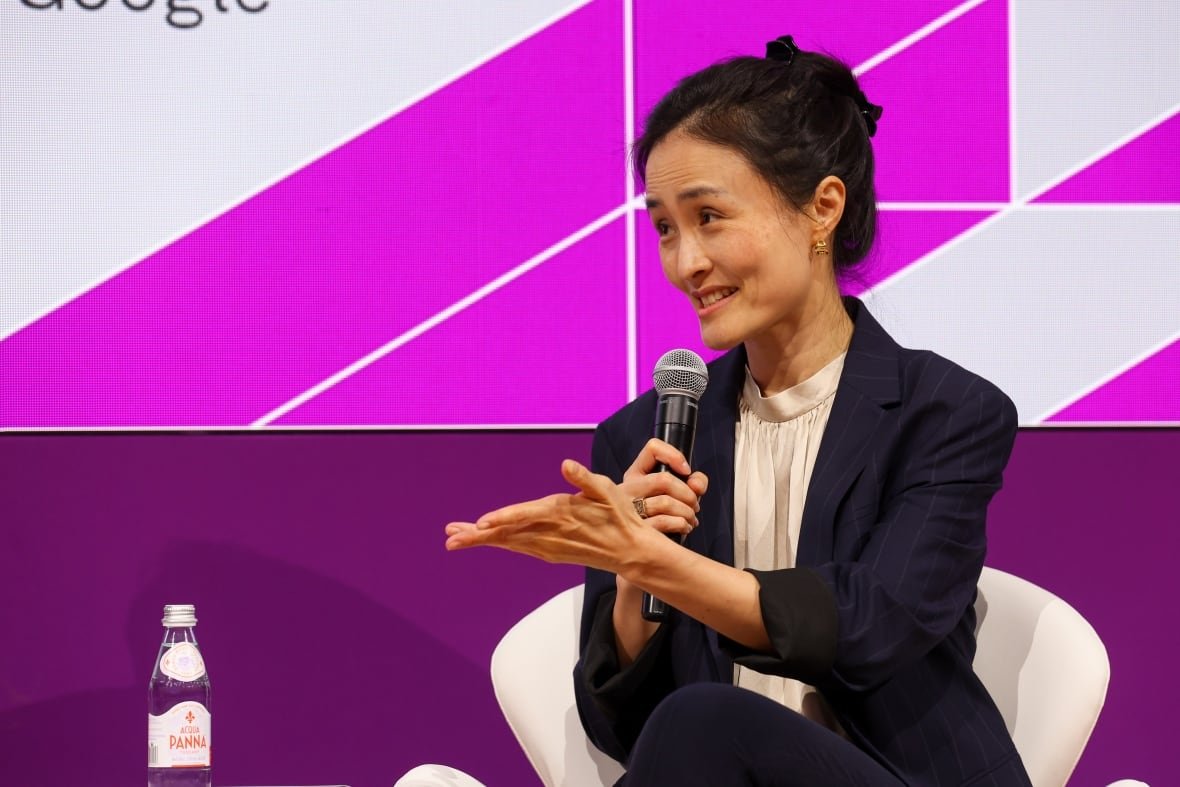 A woman wearing a blazer with her hair in a bun speaks into a microphone during a panel discussion.