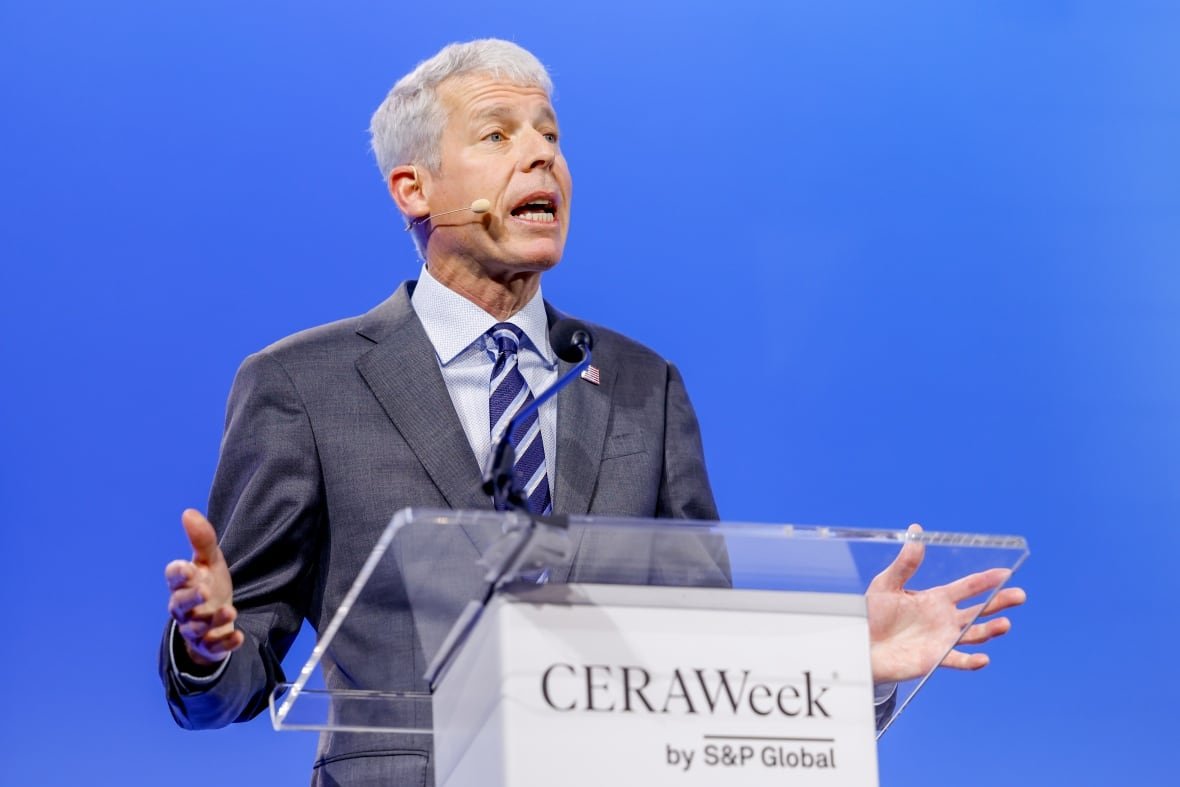 A man with white hair wearing a grey suit speaks at a podium with a blue wall behind him.