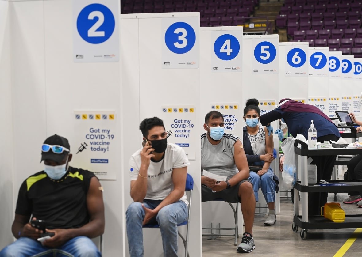 People sit in chairs, separated by dividers and wearing masks at a vaccination centre. Sleeves are rolled up and bandages visible on arms. In the back, a woman receives a vaccination from a health-care worker.