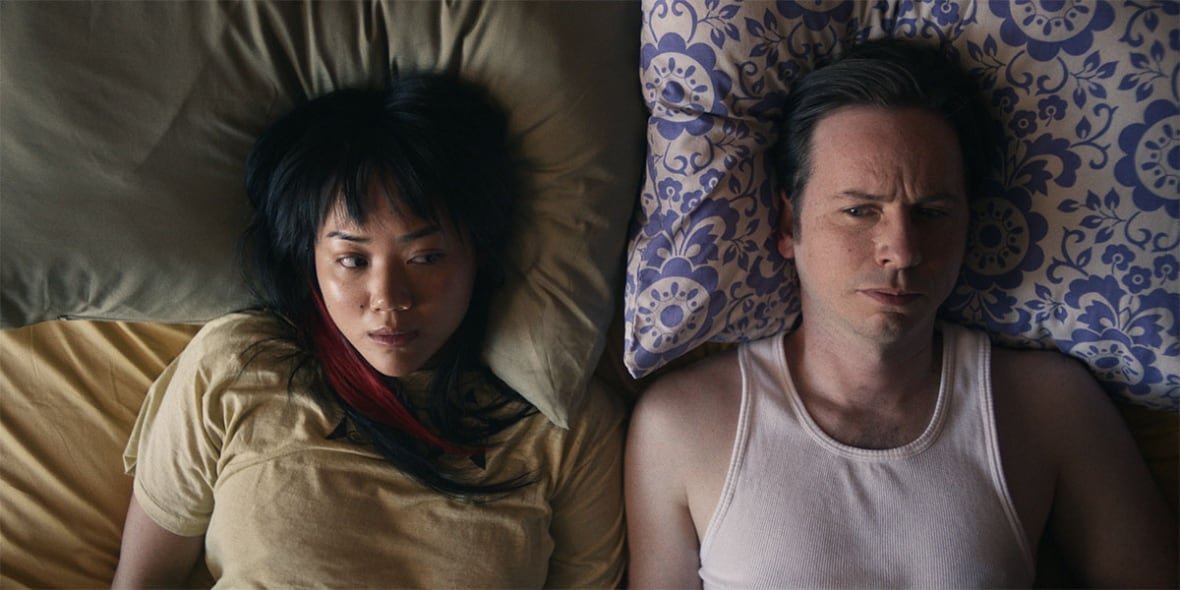 A man and woman lie next to each other in bed, eyeing each other sideways. 