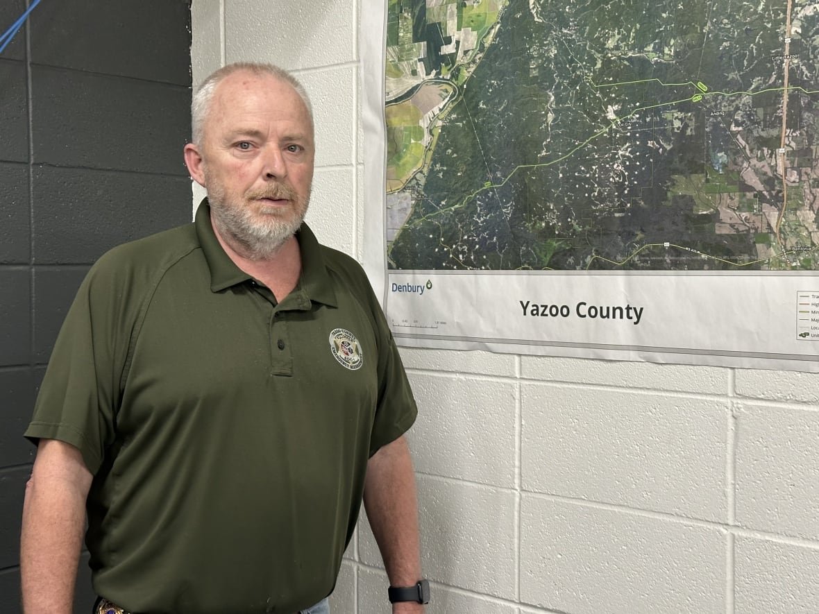 An adult white man stands in an office next to a map with the label Yazoo County.