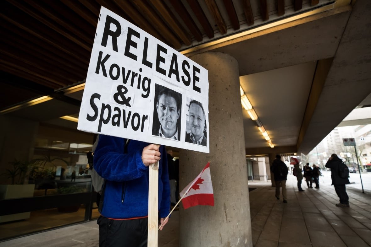 A person in a blue windbreaker holds a large sign with the black and white images of two men and text reading: Release Kovrig & Spavor. In his other hand is a small Canadian flag.