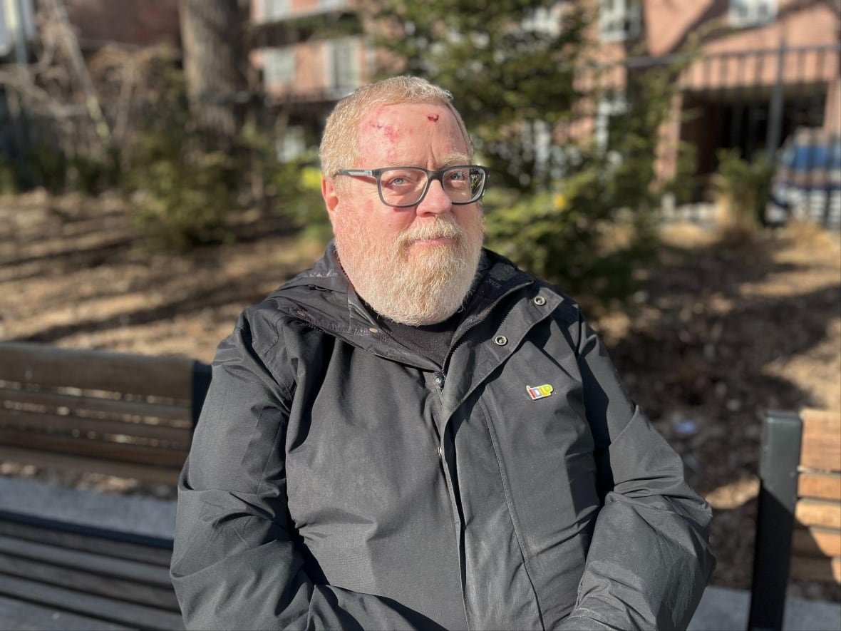 Ken Monteith, a 64-year-old man, looks into the camera, sitting on a park bench in the sunlight, wearing glasses and a light black jacket.