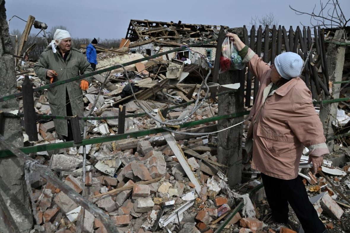 Two older women stand in the rubble of a bombed-out village. 