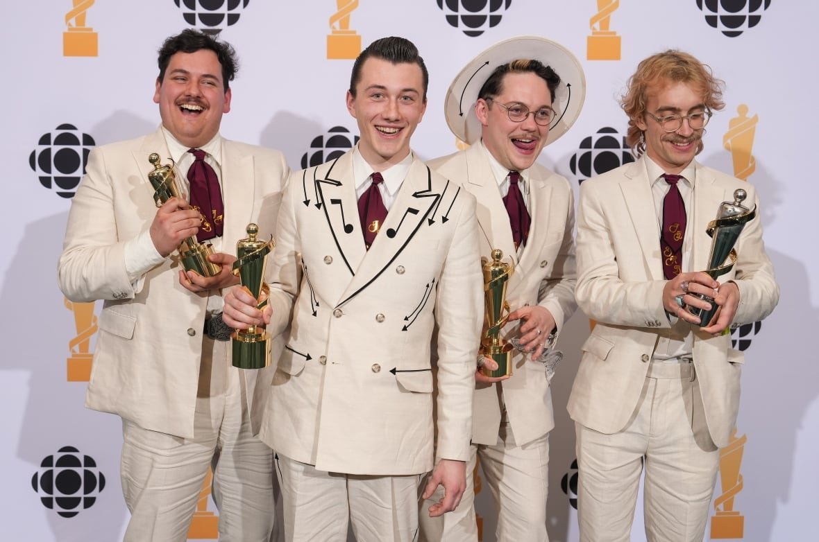 Dressed in matching retro suits with music note details on the collars, four musicians celebrating with their trophy after winning a big musical award. 