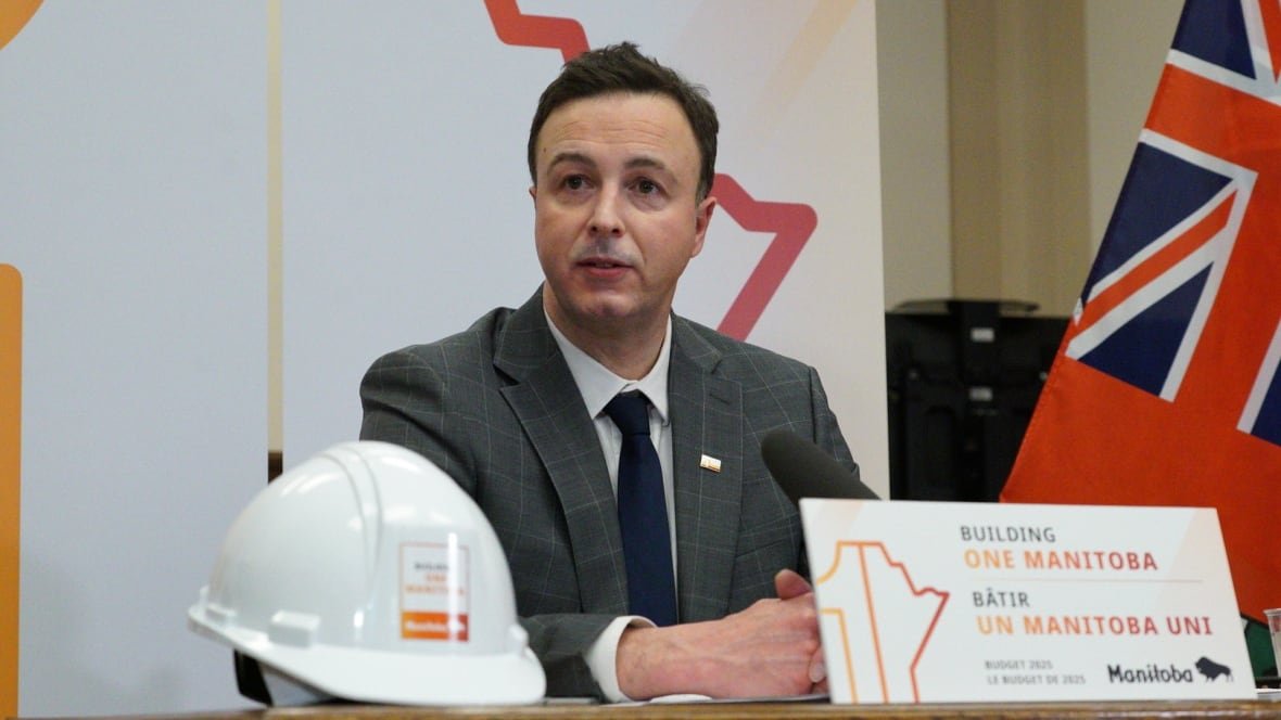 A man in a grey suits sits at a table. A white hardhat sits on the table in front of him, along with a sign reading "Building One Manitoba."