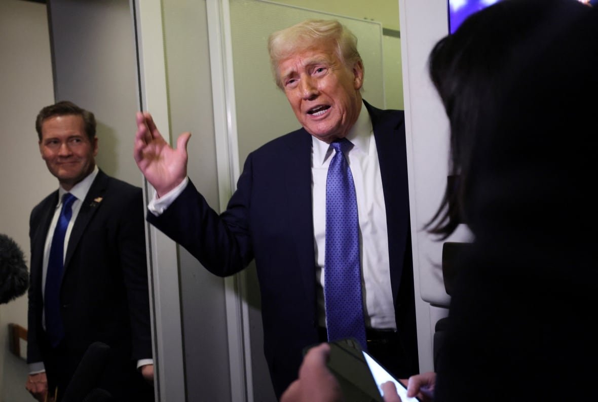 Two clean shaven men in suit and tie are shown standing. The older, light haired man is speaking while the darker haired man looks on.