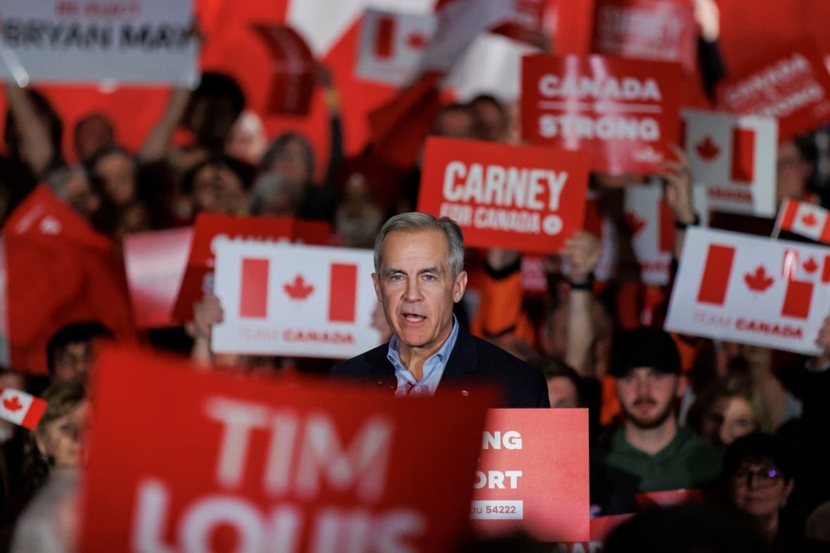 A man speaks at a podium while surrounded by a crowd carrying signs and flags.