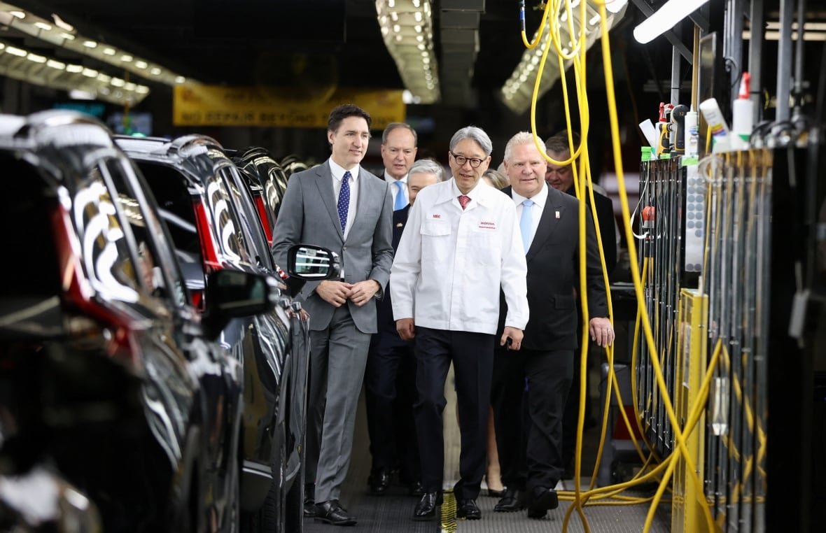 Three men walking inside a car assembly plant, with vehicles on one side and equipment on the other.