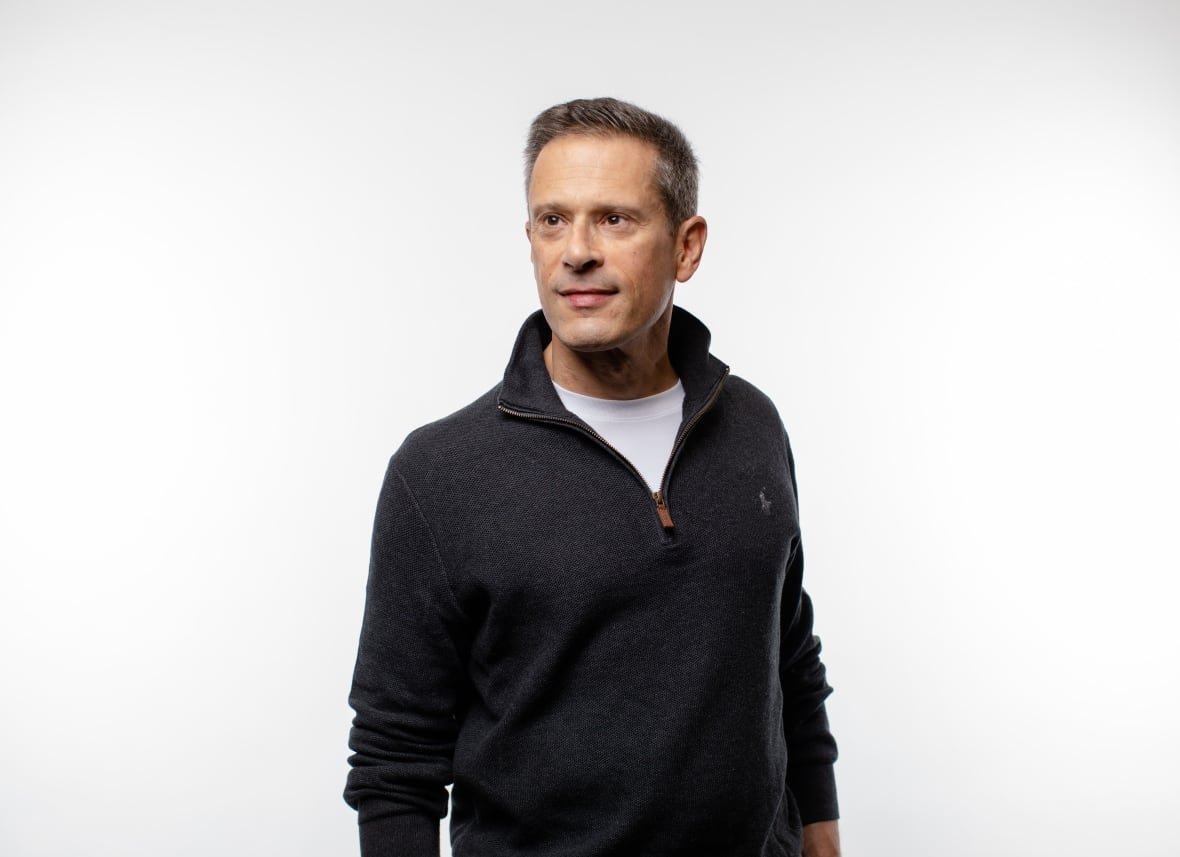 A man wearing a sweater poses in front of a white background. 