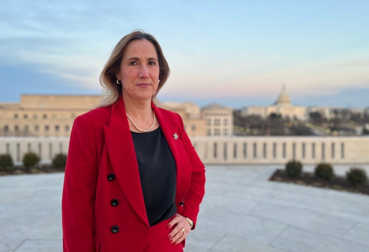 A woman with brown hair, wearing a red suit, stands near the U.S Capitol building.