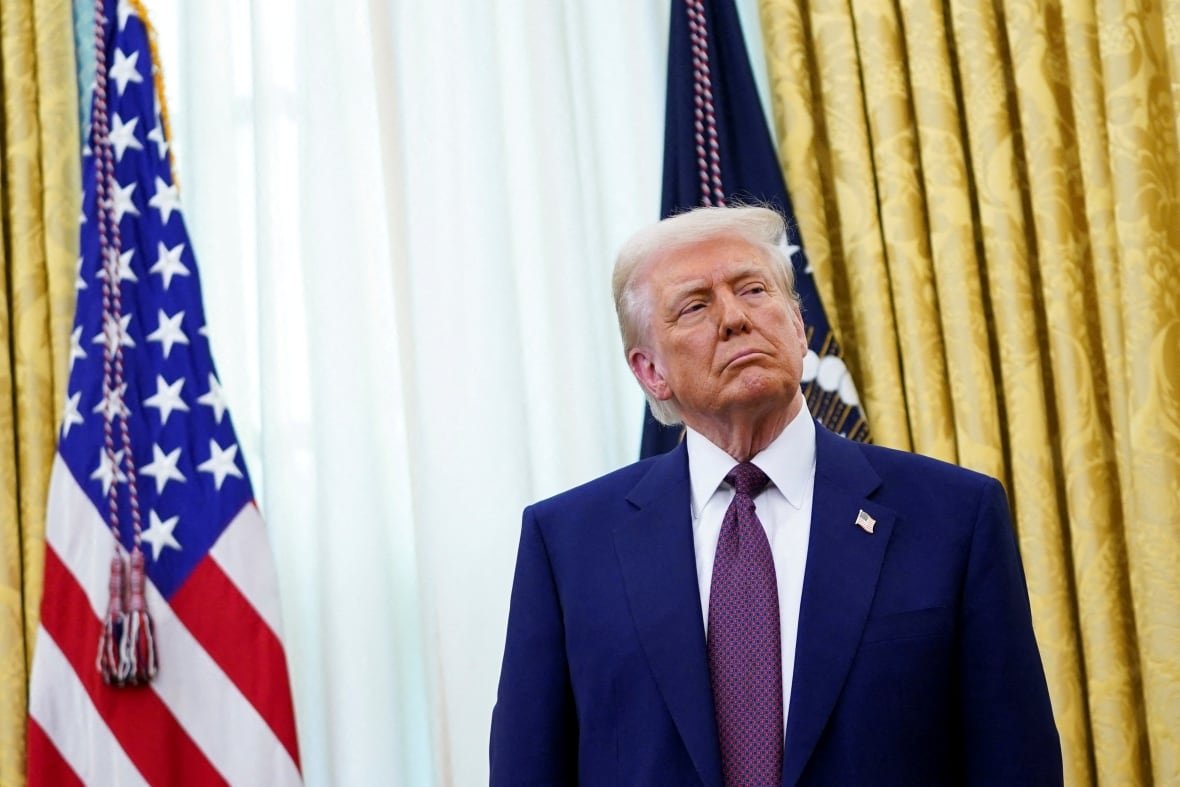 A man in a navy suit with a purple tie stands in an office with American flags behind him.