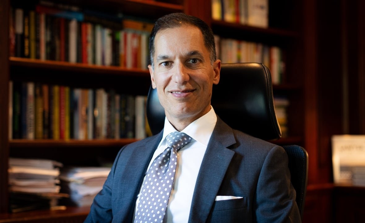 A man in a dark suit and a blue polka dot tie smiles while seated in front of a book shelf. 