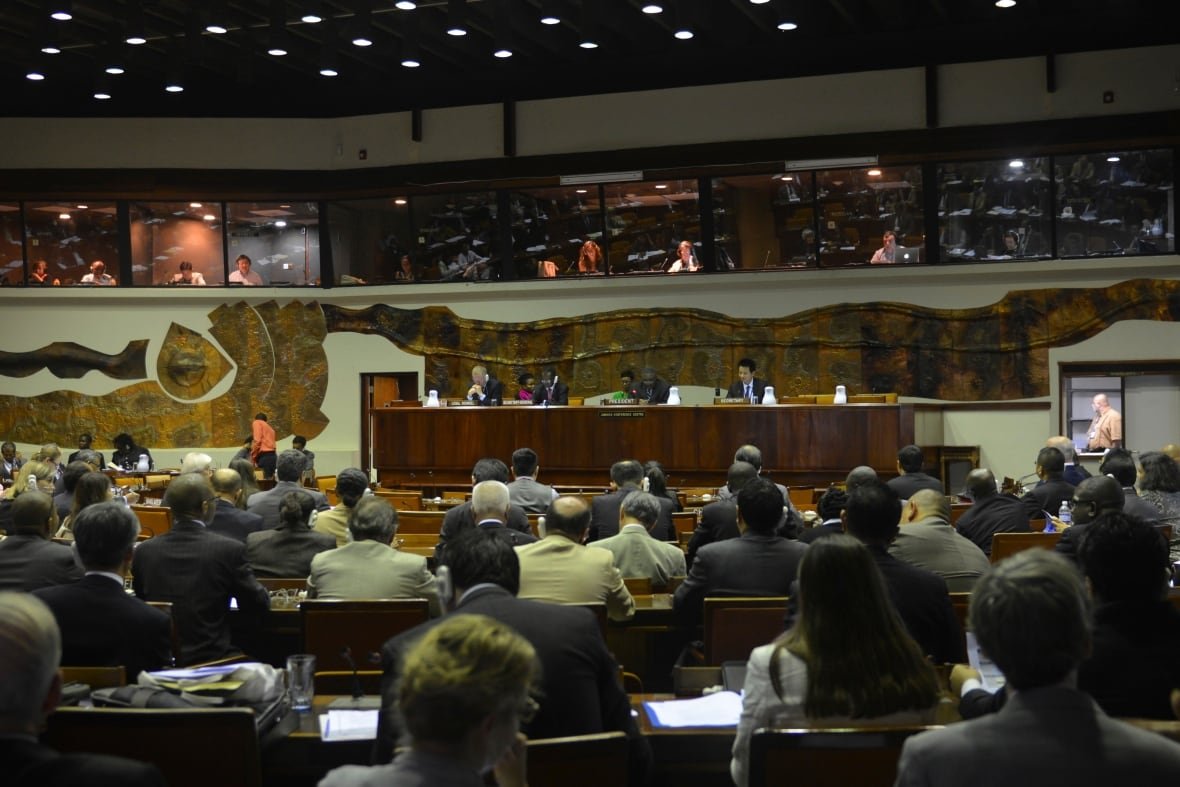 Men and women seated in a large meeting hall sit looking toward a stage and a large conference table. 