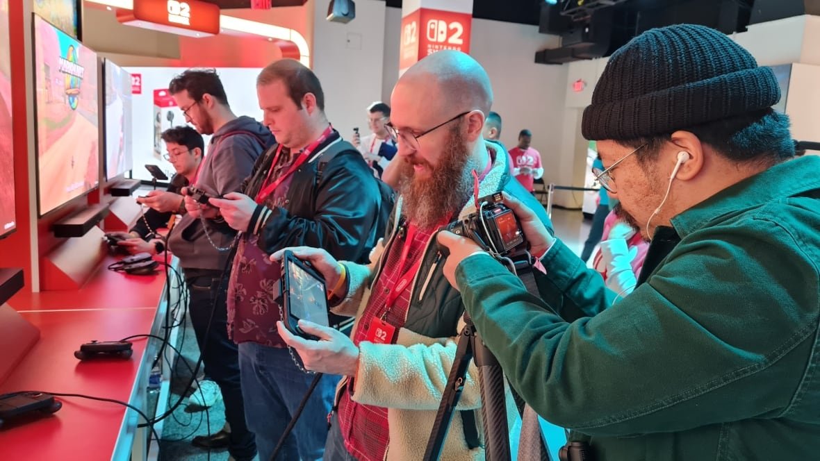 A group of men stand in a line playing a handheld videogame while another man points a camera at them. 
