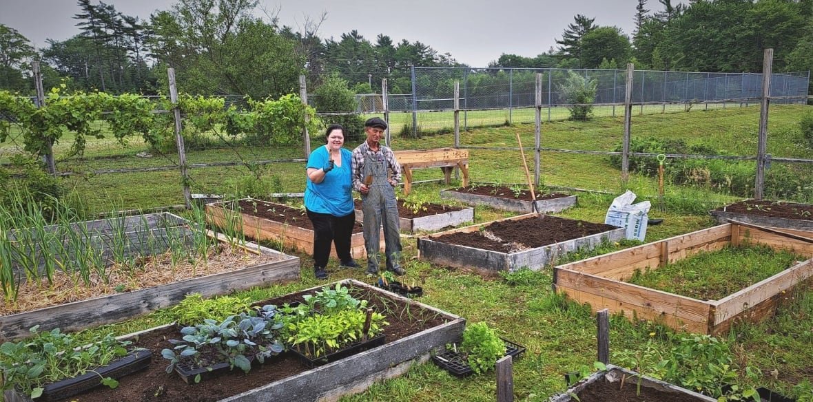 Two people are standing outside between several small garden beds. One woman wears a blue shirt, a man wears overalls and a plaid shirt. 