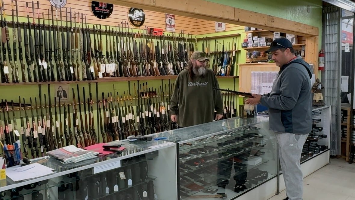 A man stands next to a counter full of knives. He's holding a rifle in his hands. An employee behind the counter looks on. 