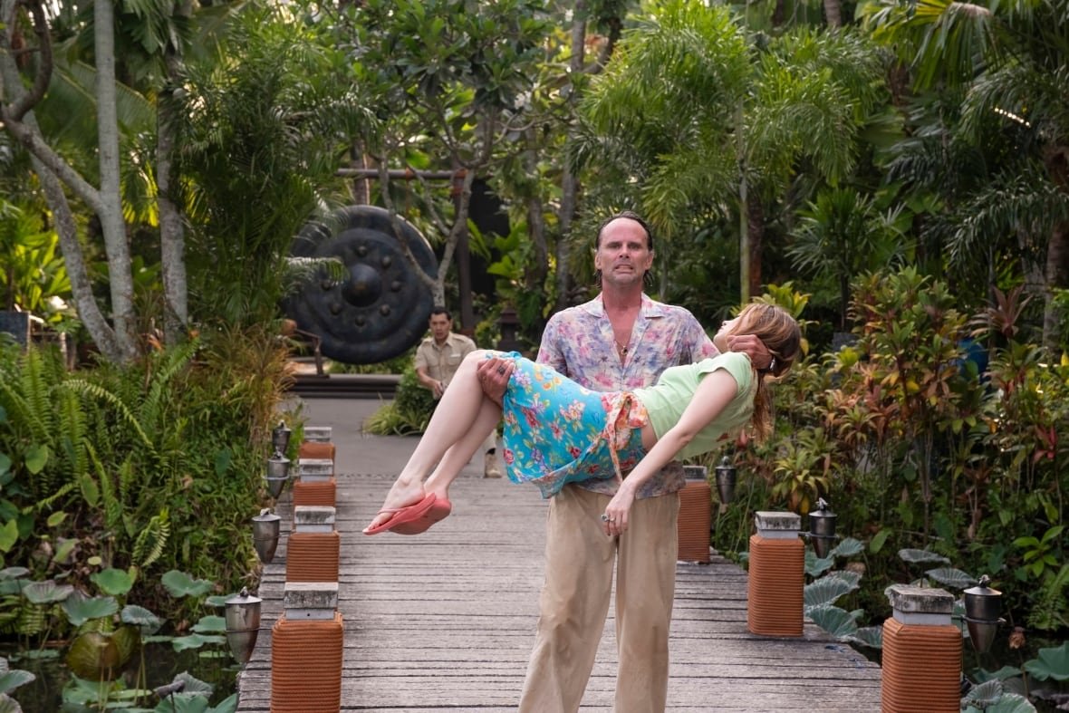 A man carries an unconscious woman in his arms. Kneeling far behind him is a man in a security guard outfit. 