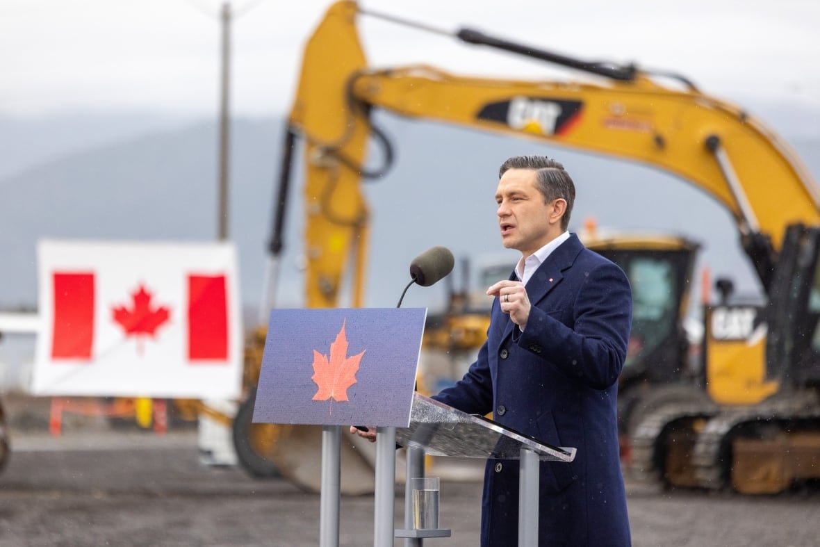 A man wearing a blue coat speaks at a podium, with a Canadian flag and front loaders behind him.