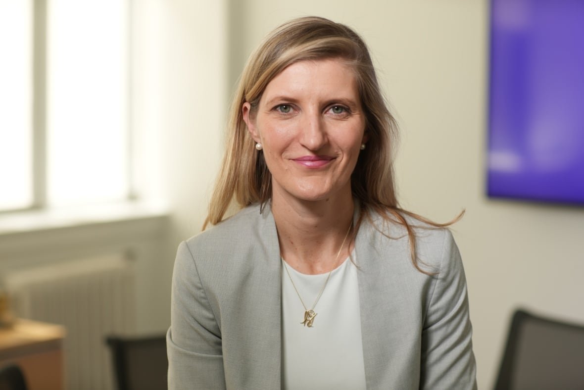 A woman with long, blond hairm wearing a light grey blazer, poses for a photo in an office.