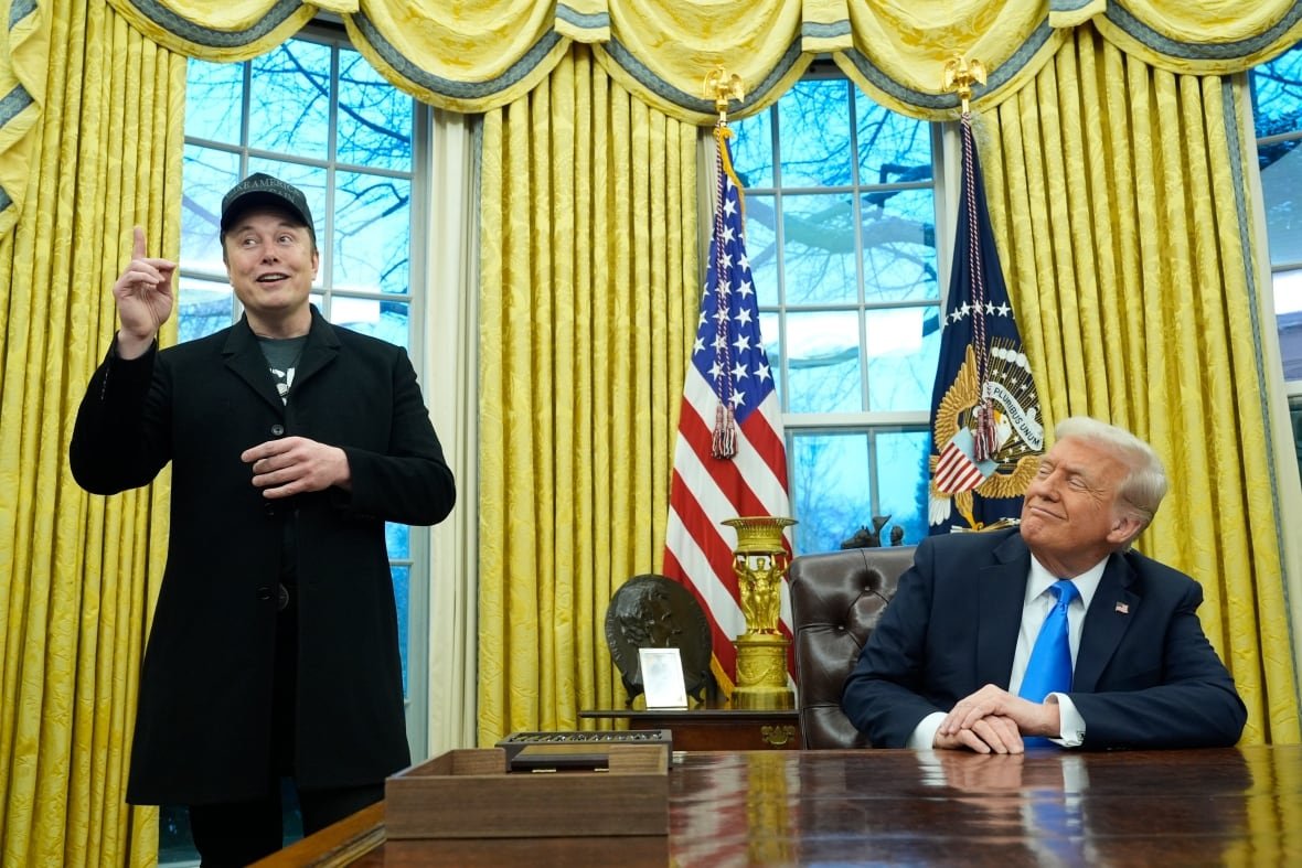 A man in a black coat and baseball cap gestures while standing next to a man in a suit seated at a desk. Yellow curtains and two flags are shown in the background.