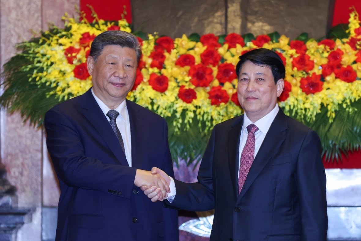 Two men in dark suits shake hands in front of a floral display.
