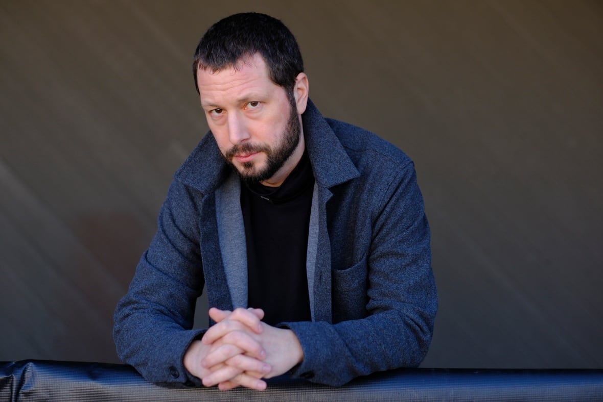 A man with short, dark hair and a beard wears a black t-shirt and grey jacket as he folds his hands together on a table, posing for a photo where he looks directly into the camera.