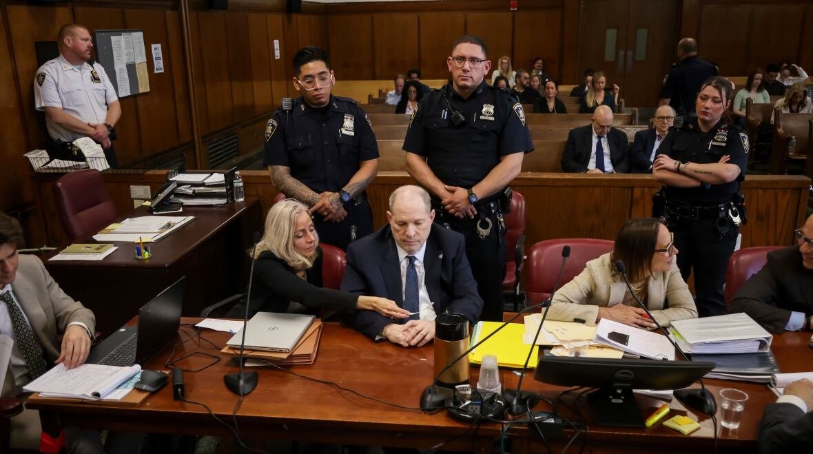 A man sits at a desk in a courtroom, with two women on either side of him, one of whom is leaning over to show him something in her hand. Two police officers stand behind the man's chair. More people are visible in the rows of seats in the courtroom at the back.