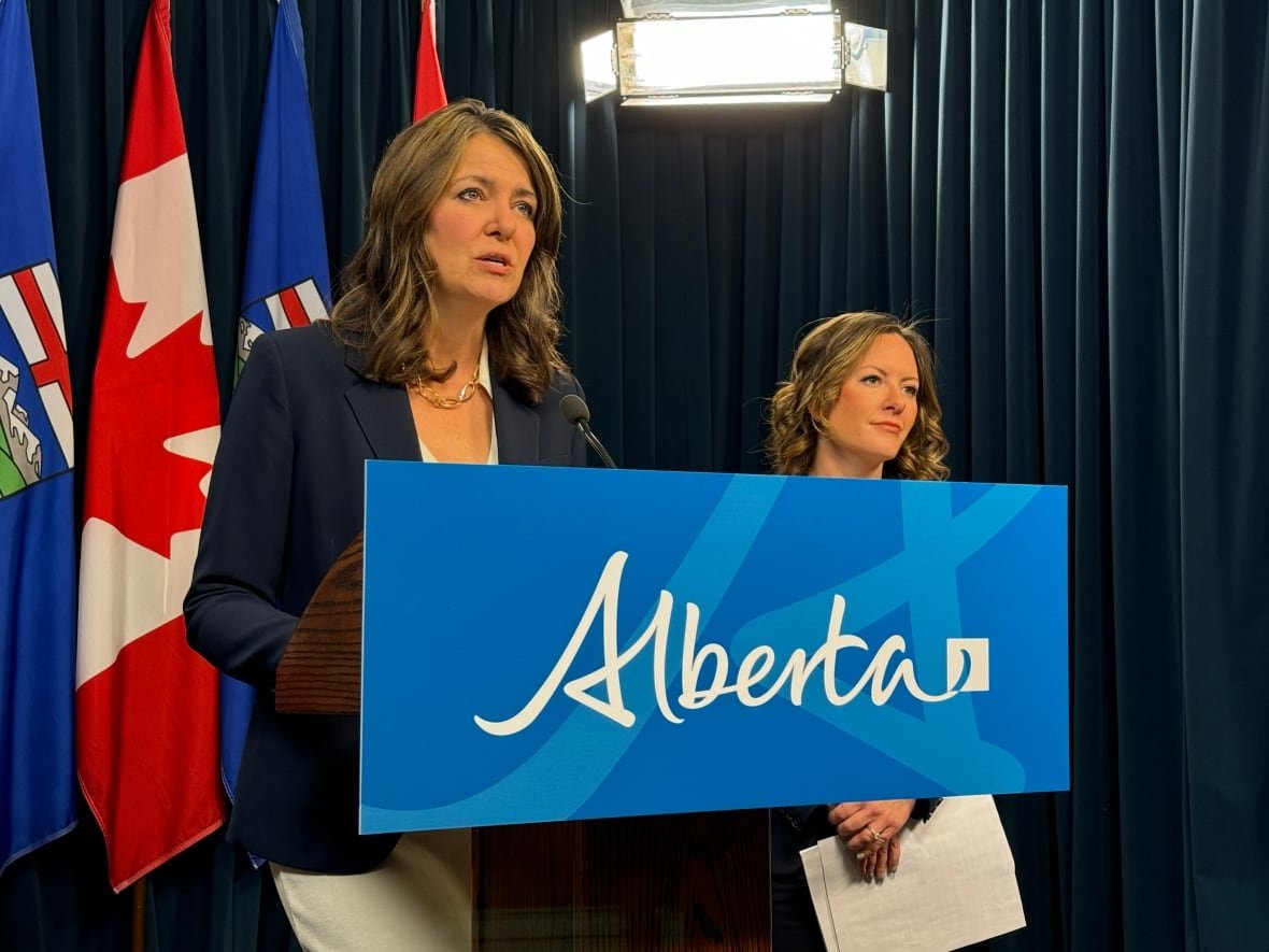 Danielle Smith, with shoulder length brown hair and wearing a navy blazer and white blouse stands in front of a lectern bearing the blue Alberta logo. Environment Minister Rebecca Schulz is beside her in the background. They are standing on a stage with dark blue curtains and Alberta and Canadian flags in the background.