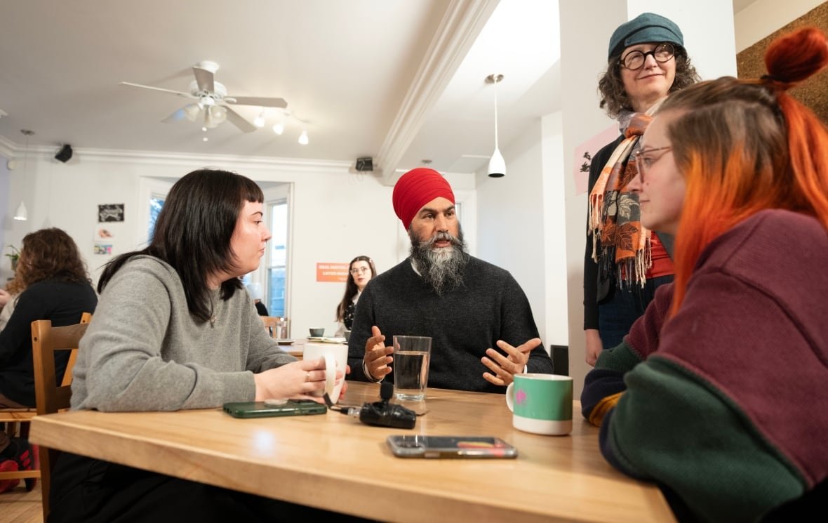 A man is seated at a table alongside two women with mugs, and another woman standing beside them.
