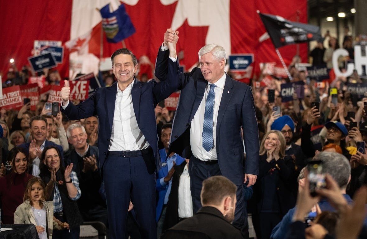 Former prime minister Stephen Harper and Conservative Party Leader Pierre Poilievre are pictured standing side-by-side on a stage, raising clasped hands. They are surrounded by a crowd of people.