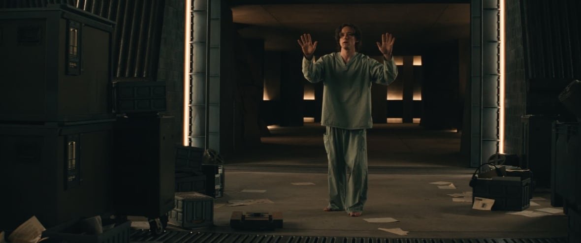 A man wearing hospital robes holds his hands in the air. He is standing in a dark room filled with papers and drawers on the ground. 