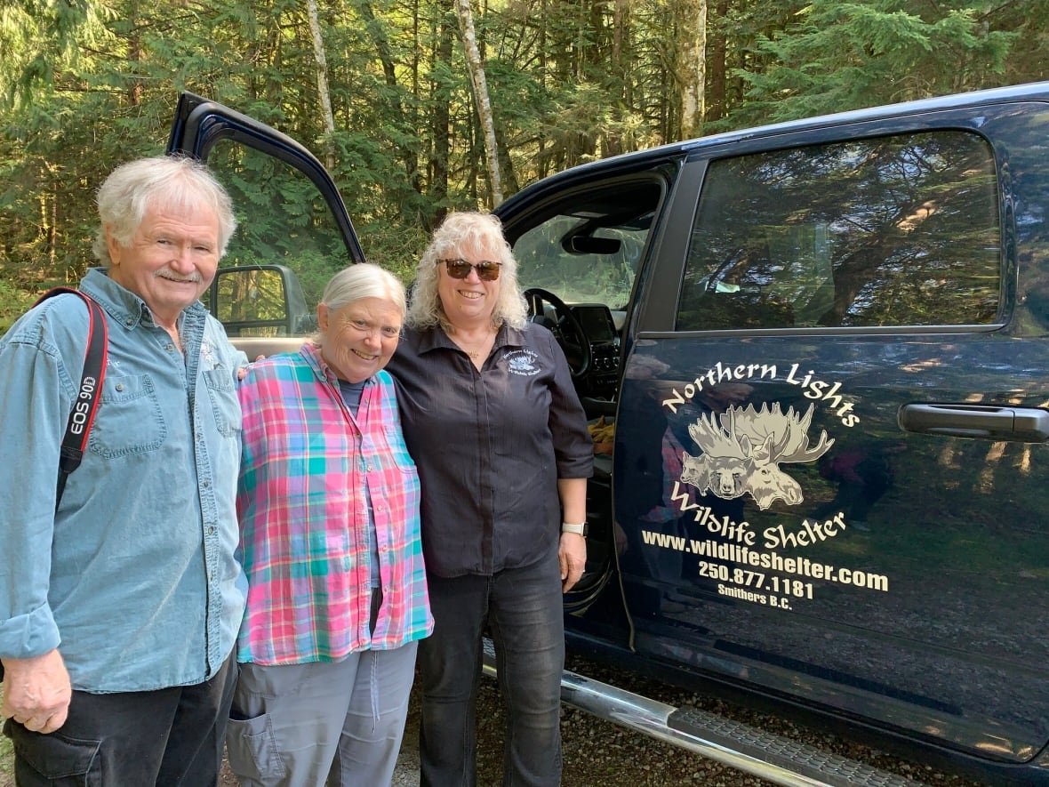Three older people pose for a picture near a black pickup truck.