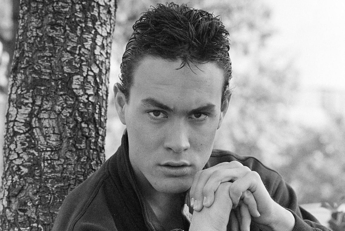 A black and white photo shows a young man looking directly at the camera, his hands folded in front of him.