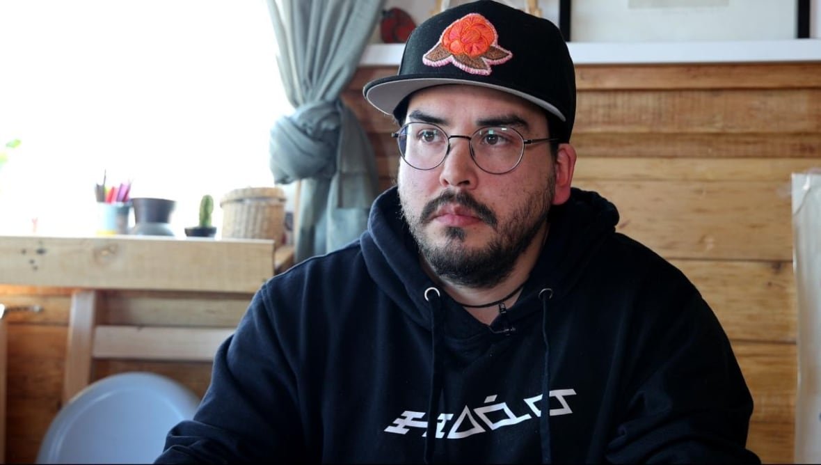 A man wearing round, black-rimmed glasses and a baseball cap sits at his kitchen table and looks on. 