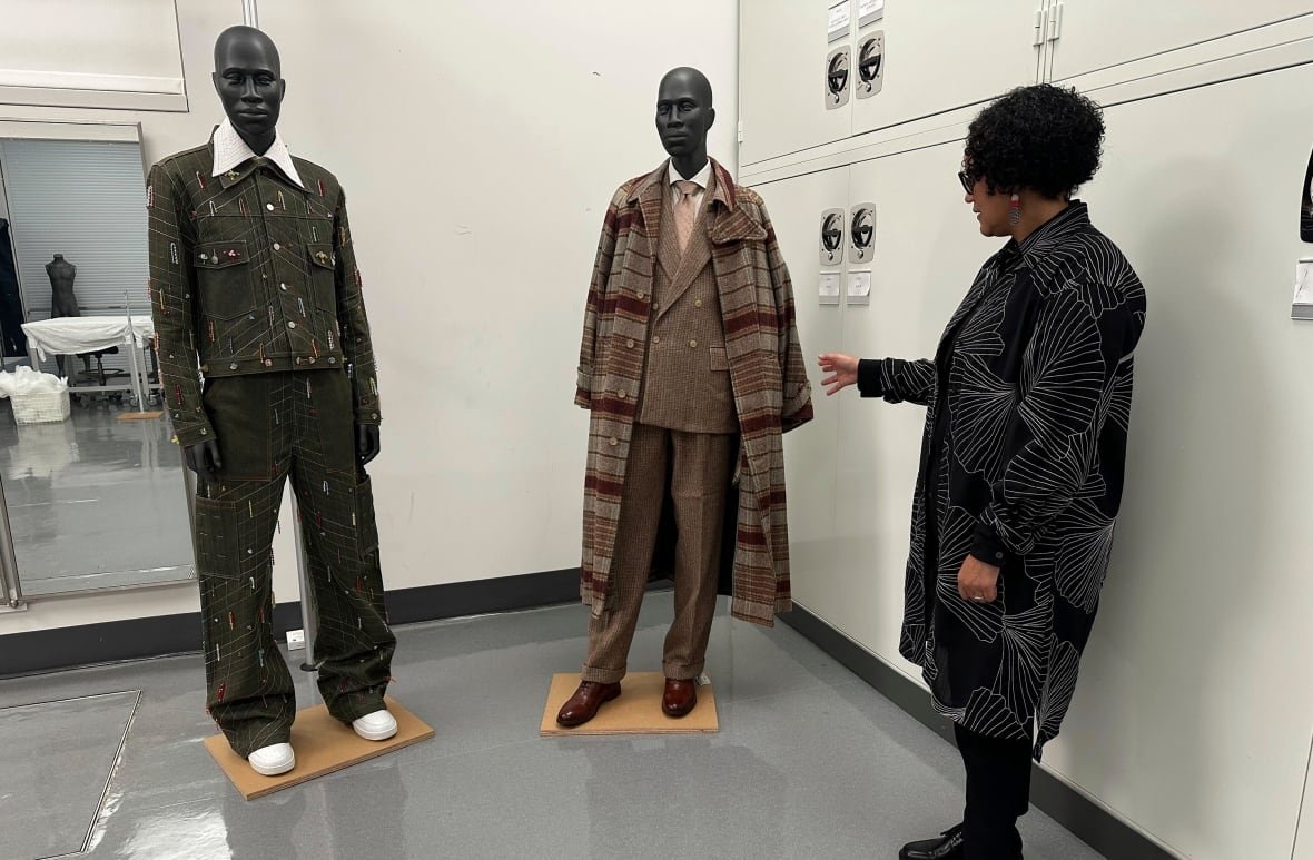 A woman gestures to two mannequins that are wearing menswear looks. One involves a long coat. 