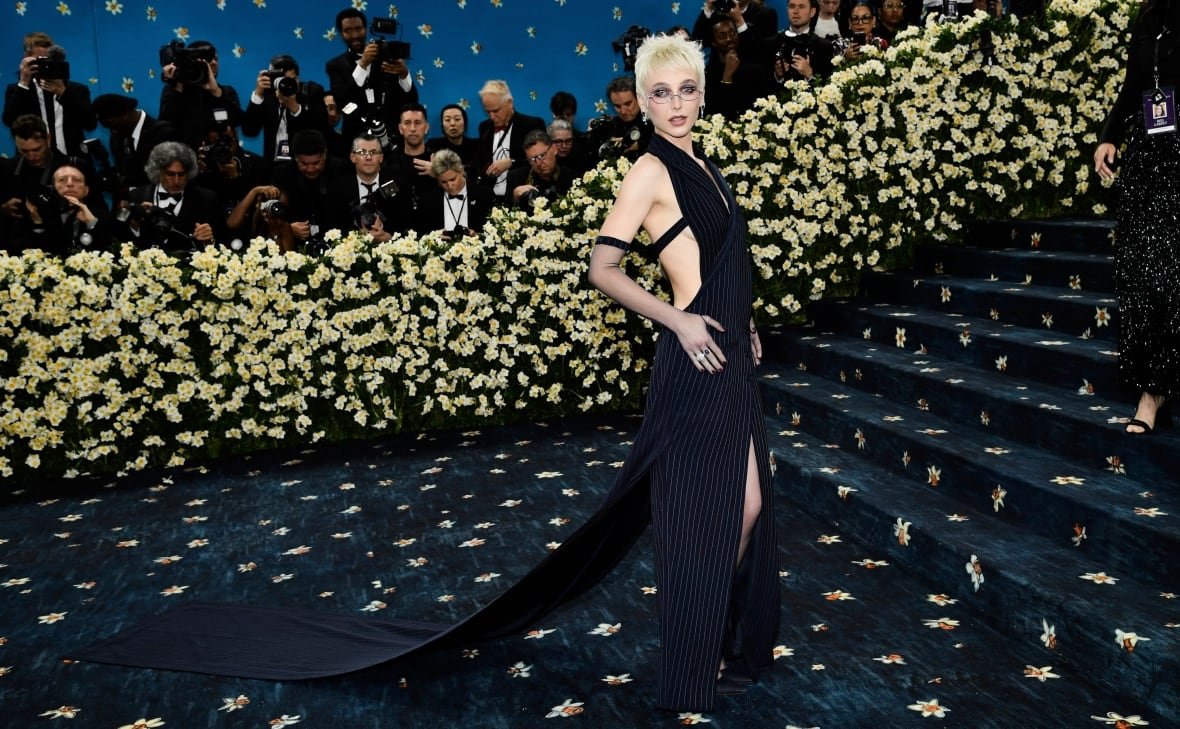 A woman with very short blonde hair poses at the bottom of a staircase in a dress that looks like a deconstructed pinstripe suit. 