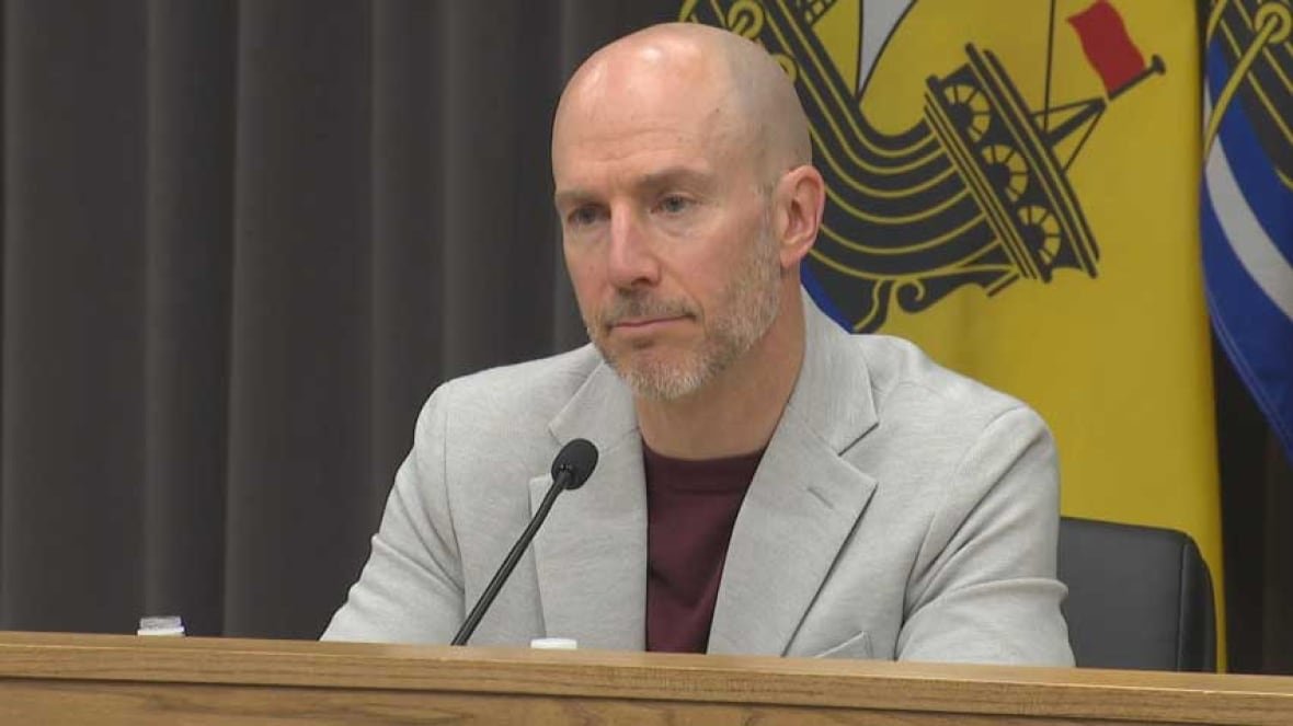A man wearing a T-shirt and blazer, sitting in front of a microphone, speaking.