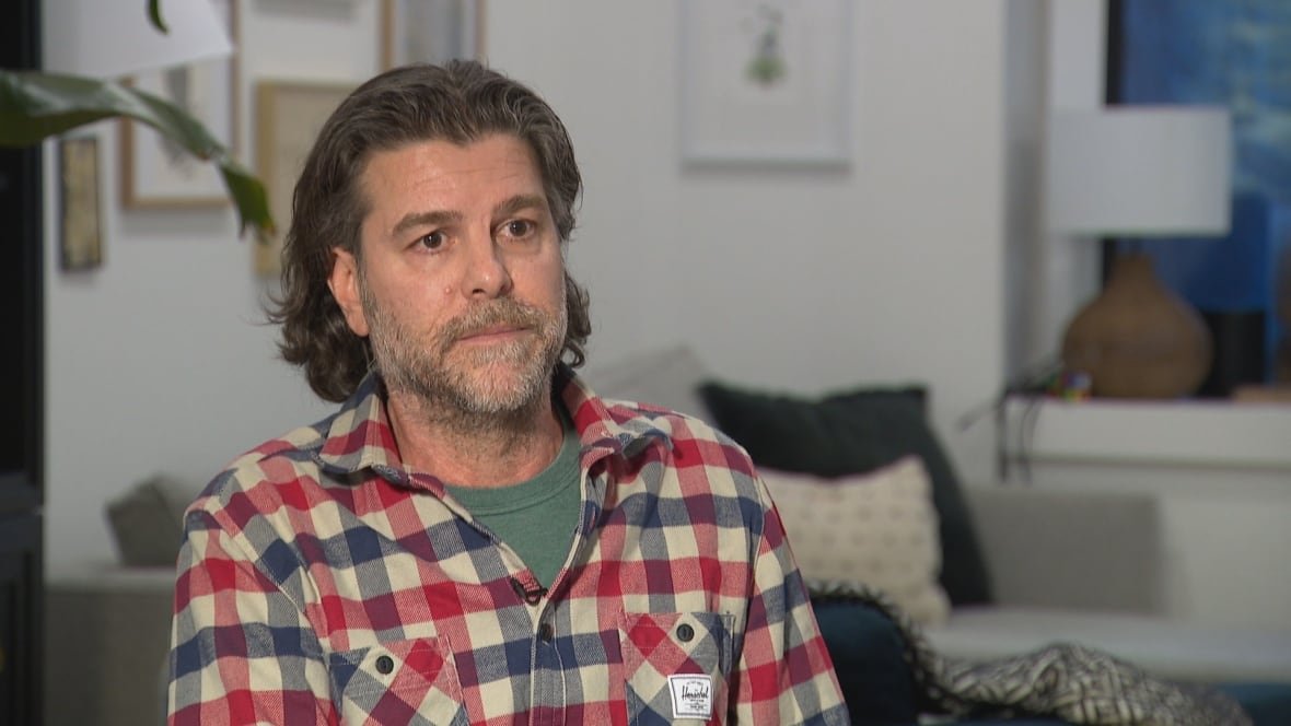 A bearded man in a red and blue checked shirt sits in an indoor living room, looking off-camera to the right.