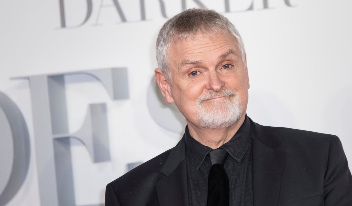 A man white white hair and a goatee is seen smiling in a black suit in front of a white backdrop. 